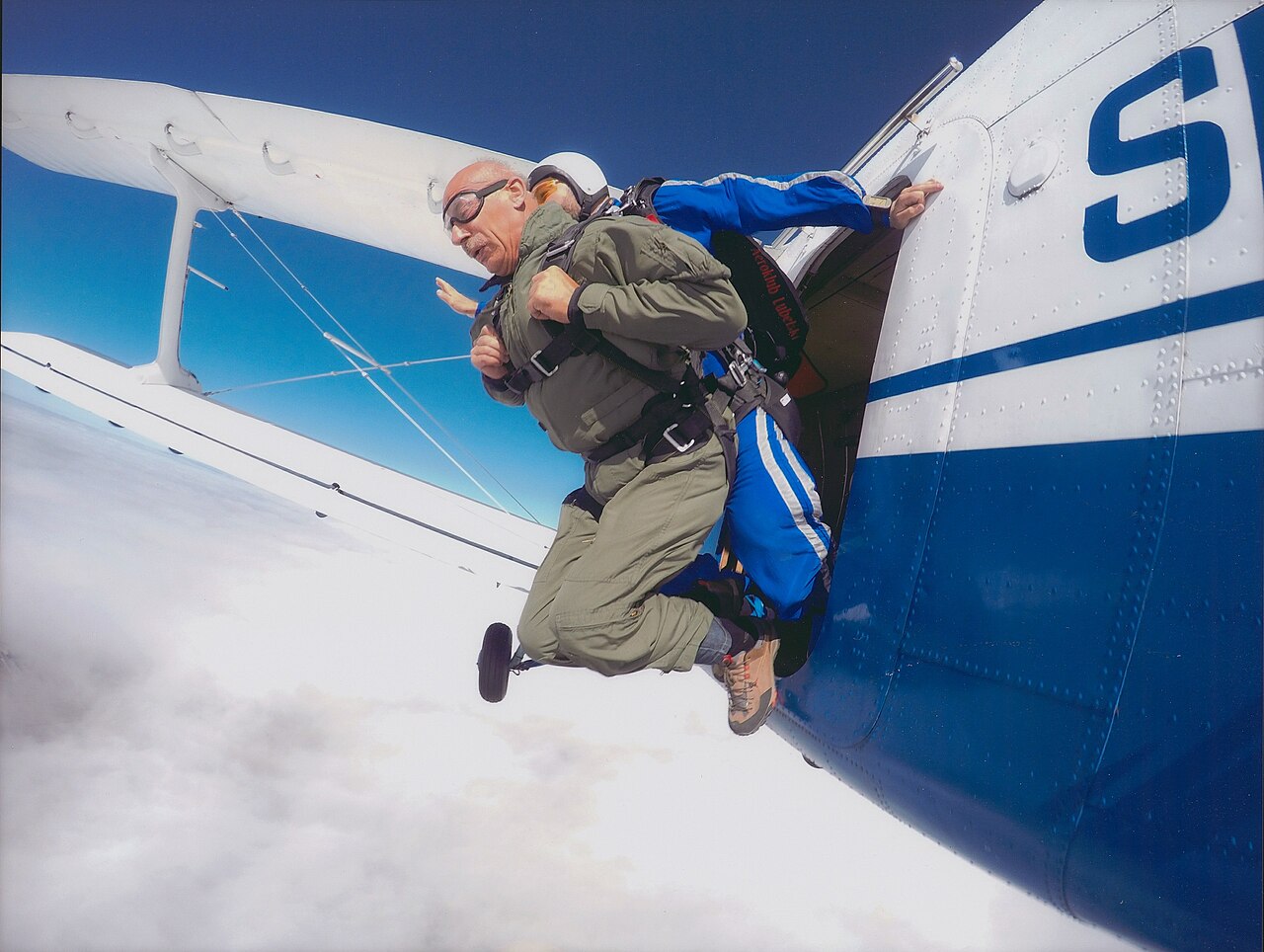 A tandem skydiving jump from an aircraft, showcasing a confident instructor and a skydiver preparing to leap into the blue sky.