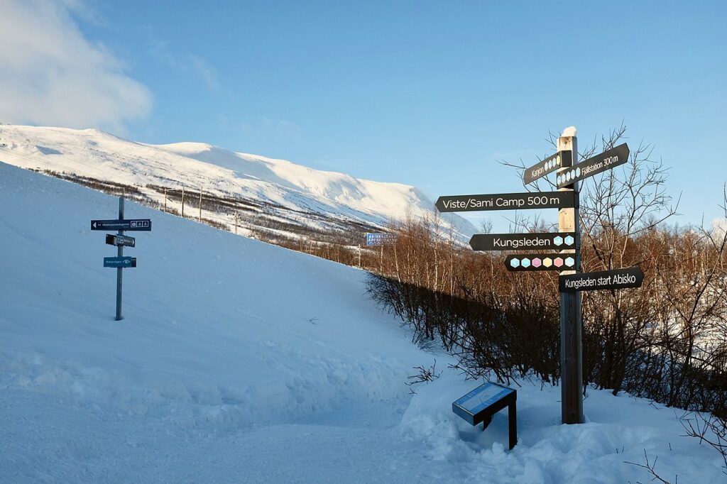 Snow-covered trail signs point towards various hiking destinations in a mountainous landscape under a clear blue sky.
