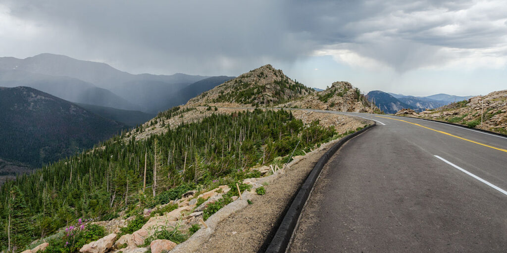 A winding road curves along a mountainside, surrounded by dense forest and distant peaks under a cloudy sky, suggesting impending rain.