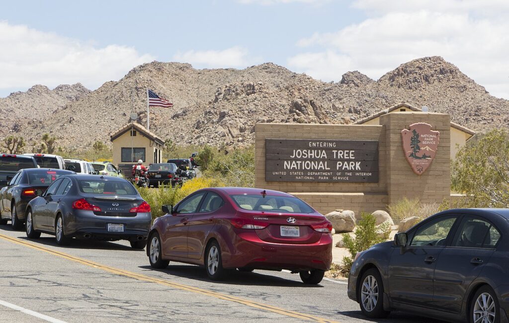 Cars lined up at the entrance of Joshua Tree National Park, with a park sign, a visitor center, and mountains in the background.