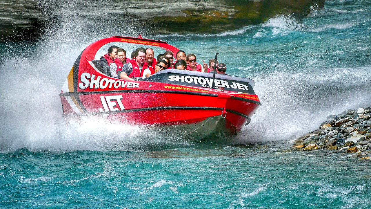 A bright red jet boat splashes through turquoise waters, surrounded by rocky riverbanks, with passengers enjoying the thrilling ride.