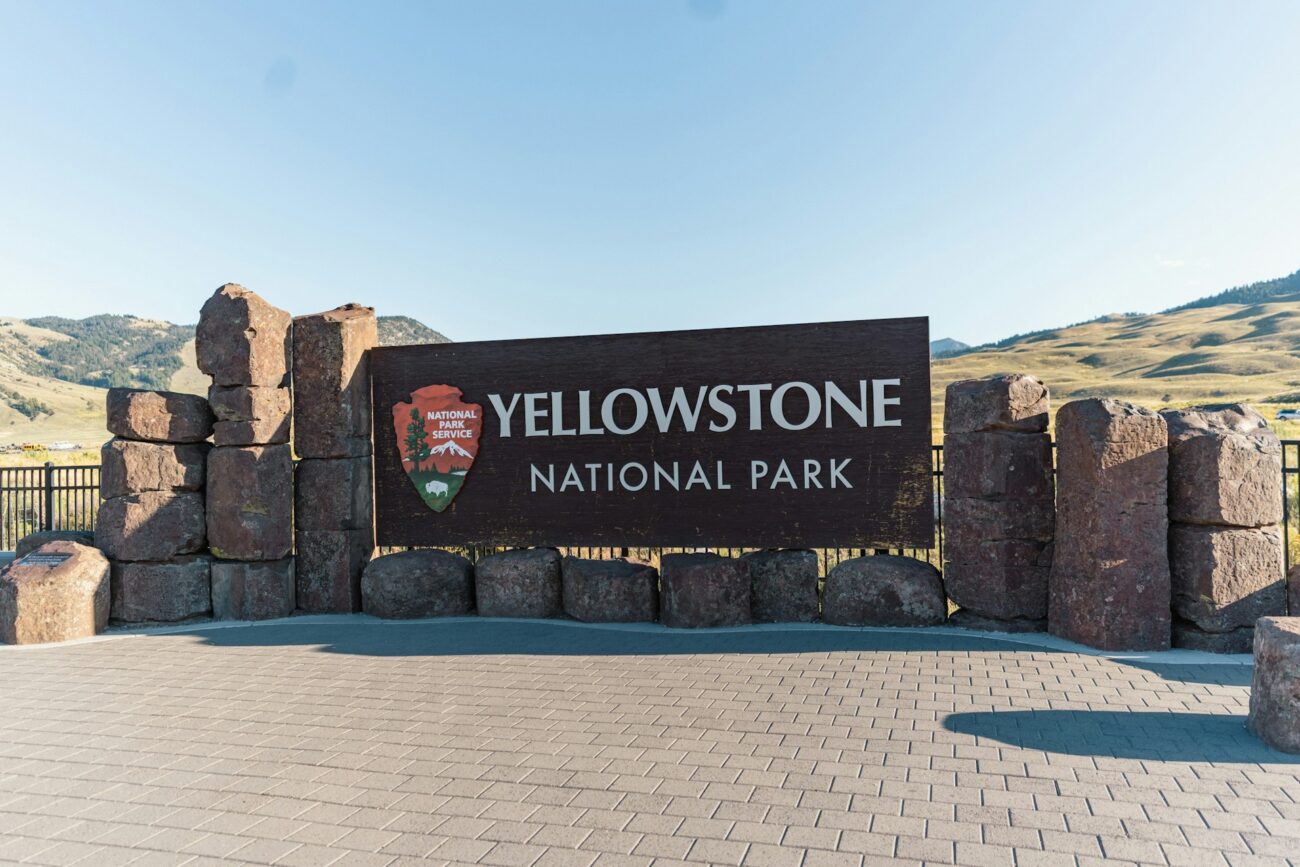 Sign welcoming visitors to Yellowstone National Park, surrounded by stone features, with mountains in the background and clear blue skies.