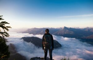 Hiker standing on a mountain peak overlooking a sea of clouds and distant mountains under a clear blue sky.