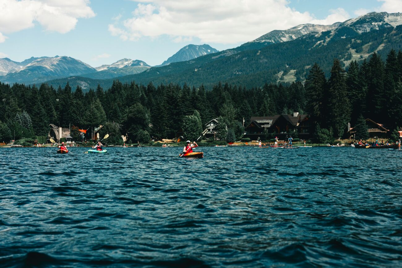 Two kayakers paddle on a serene lake surrounded by lush forests and mountains, with people enjoying various water activities nearby.