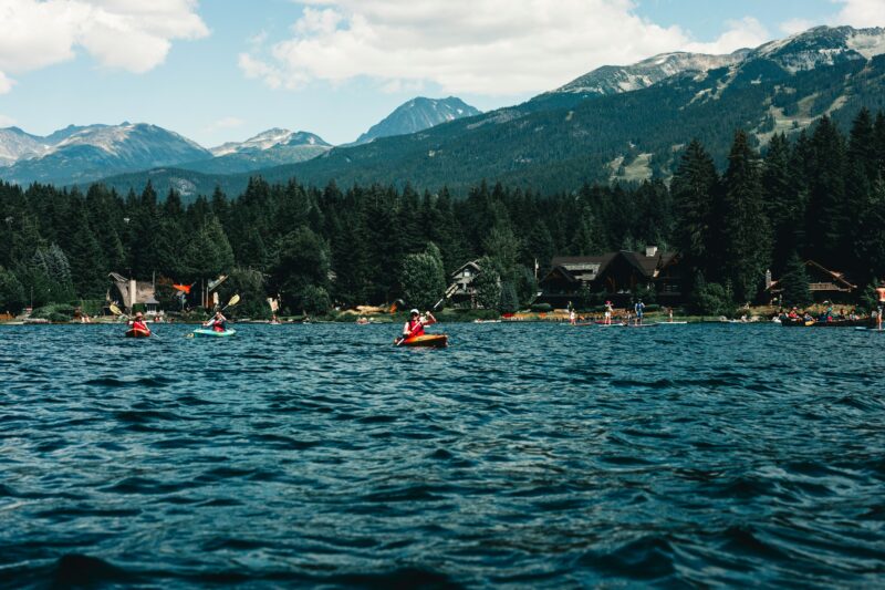 Two kayakers paddle on a serene lake surrounded by lush forests and mountains, with people enjoying various water activities nearby.