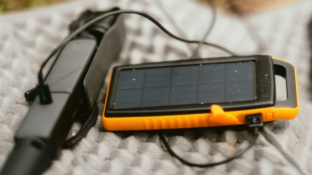 A close-up of a portable solar charger and a device, connected by a cable, resting on a textured surface outdoors.