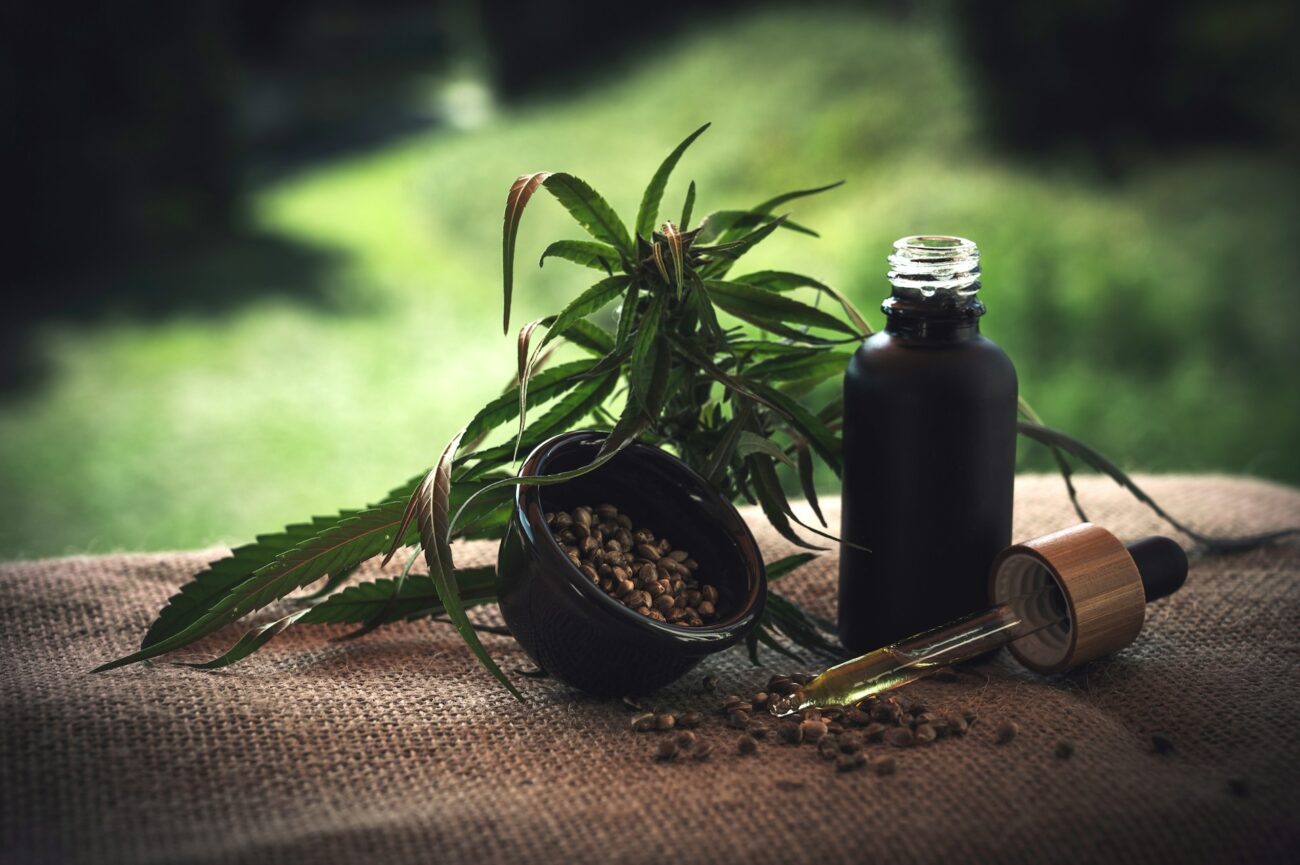 A black dropper bottle and a bowl of hemp seeds are placed on burlap, surrounded by green hemp leaves outdoors.