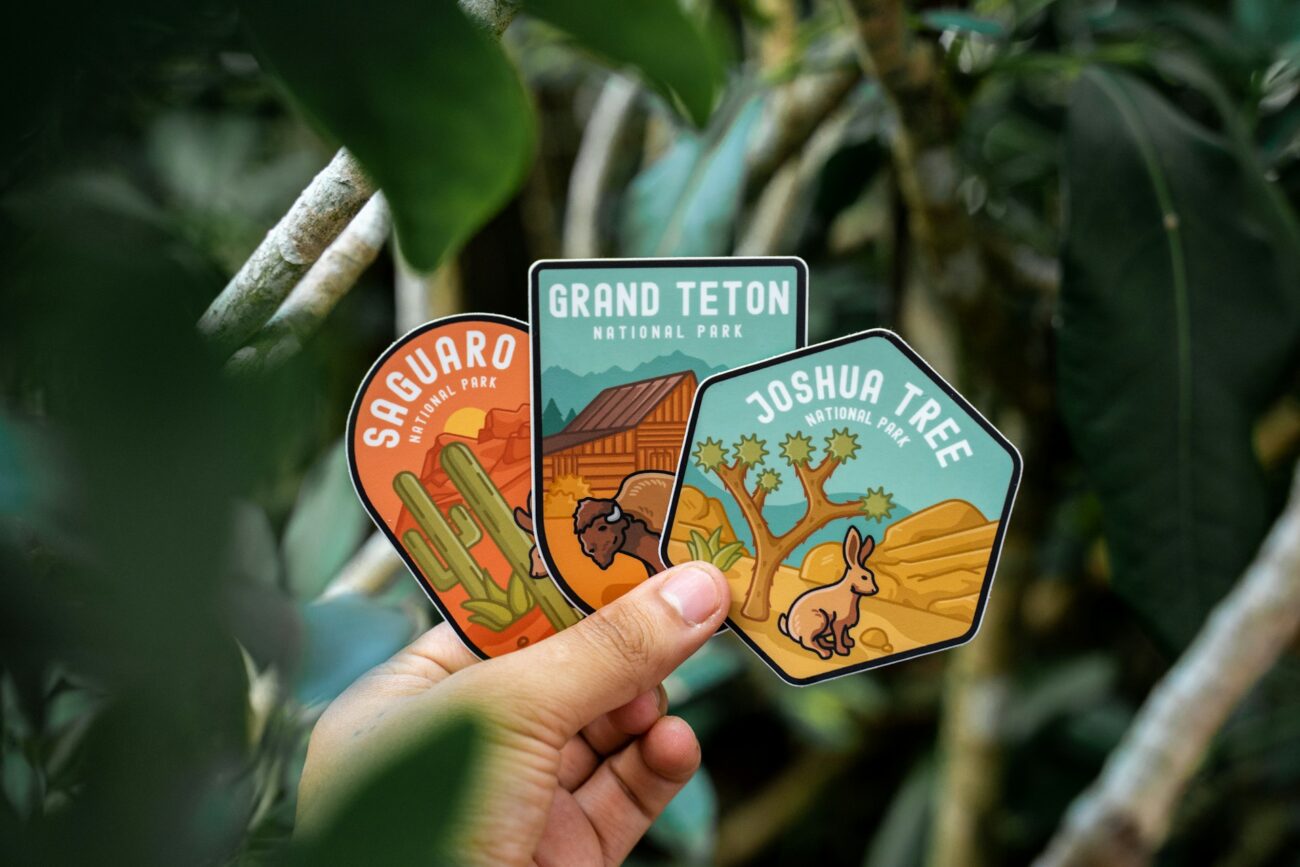 A hand holds three colorful national park stickers: Saguaro, Grand Teton, and Joshua Tree, surrounded by lush green foliage.