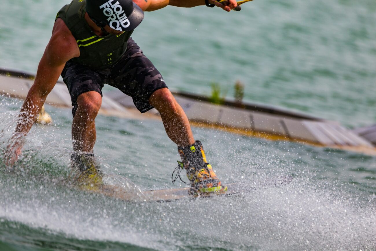 A wakeboarder performs a trick on the water, splashing as he carves sharply, wearing a life vest and helmet at a wake park.