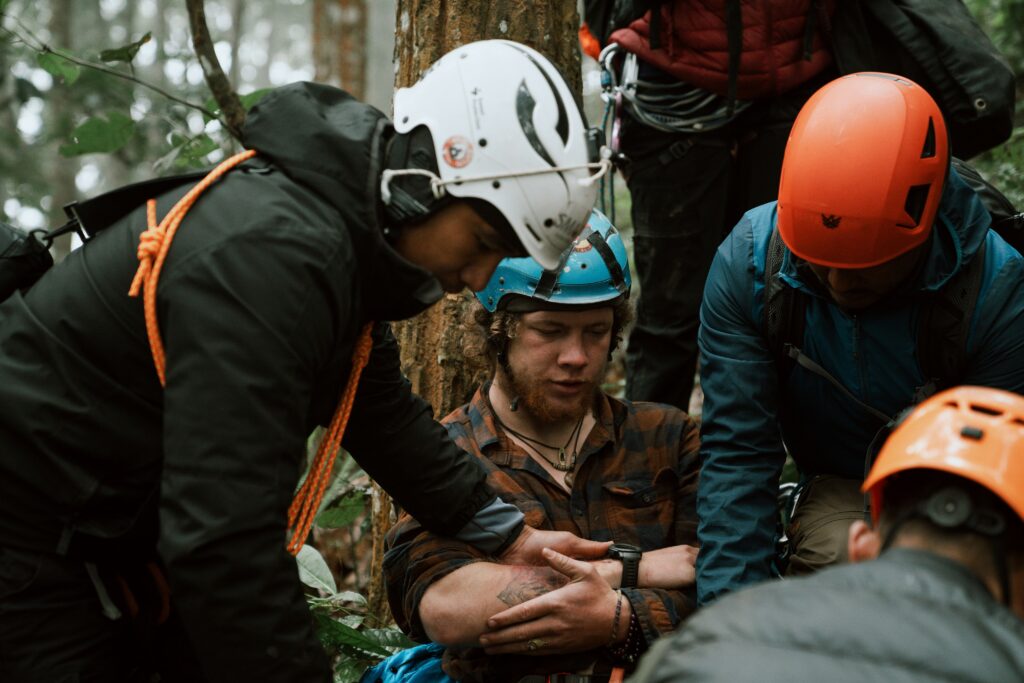 Rescue team assists an injured climber in a forest, surrounded by trees and rocky terrain, focused on providing support and care.