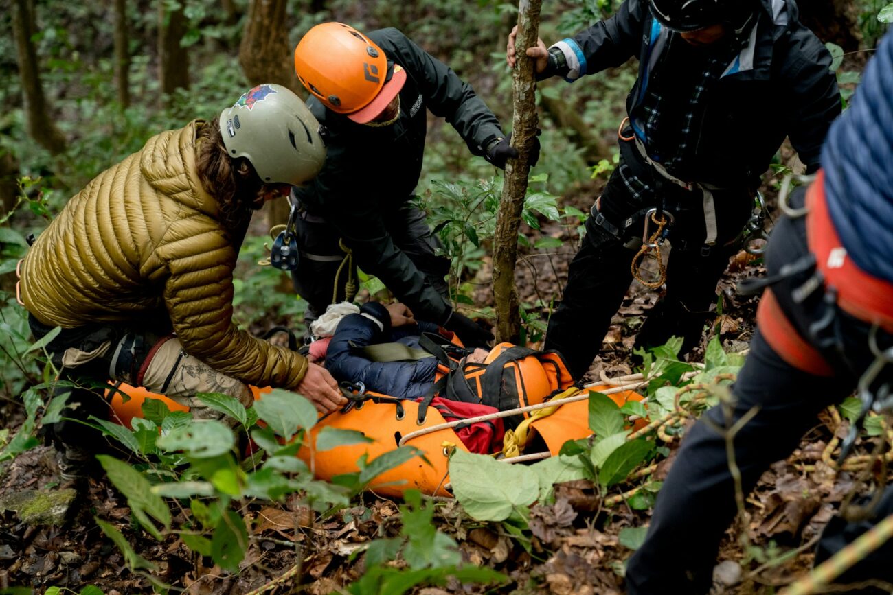 Rescue team members assist an injured climber on a steep forest slope, using a bright orange stretcher surrounded by dense foliage.