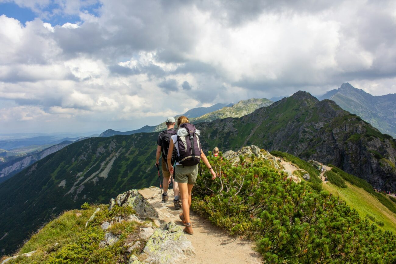 Two hikers traverse a narrow mountain path, surrounded by lush green hills and a dramatic cloudy sky.
