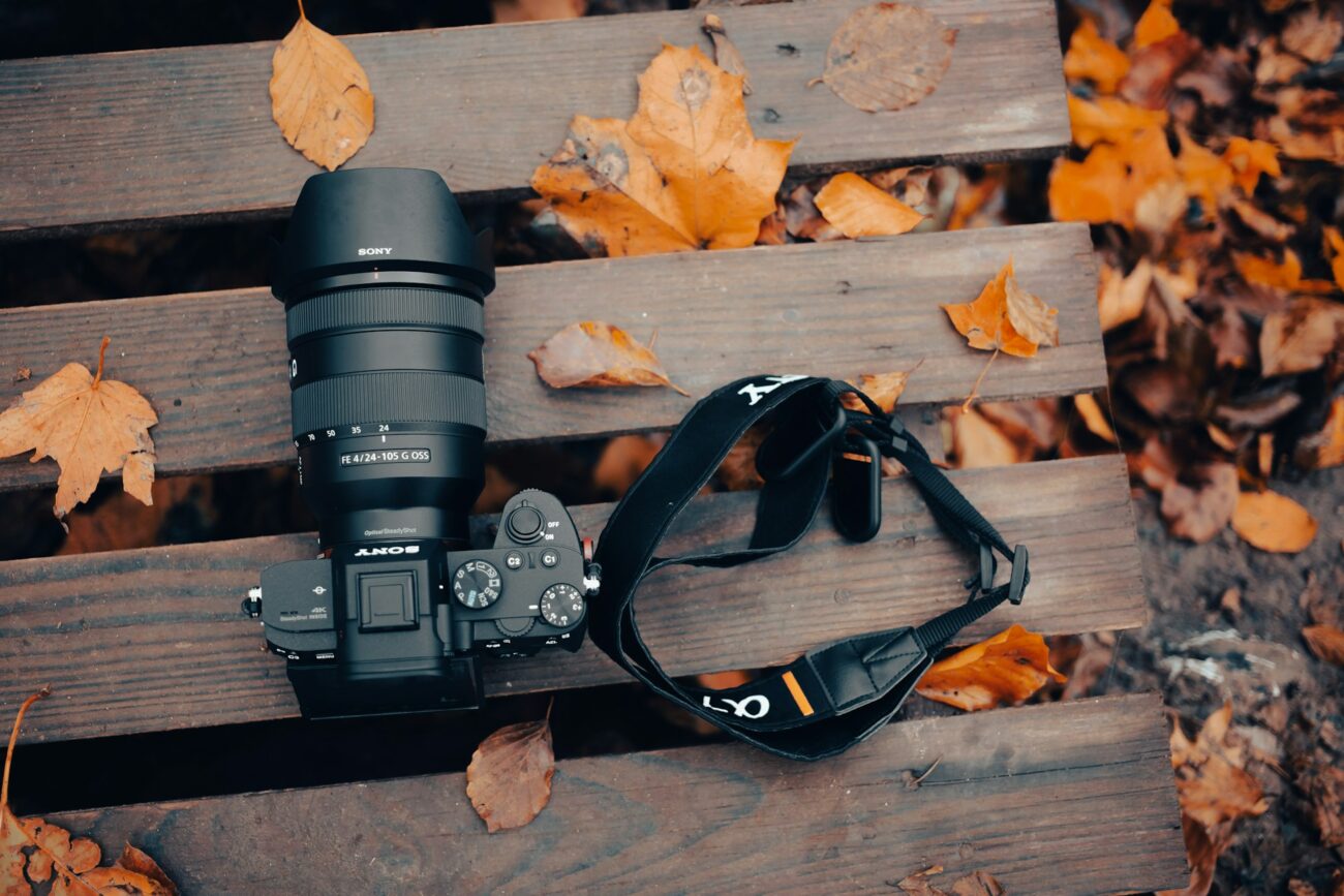 A Sony camera with a 24-105mm lens rests on wooden planks scattered with autumn leaves, accompanied by a black camera strap.