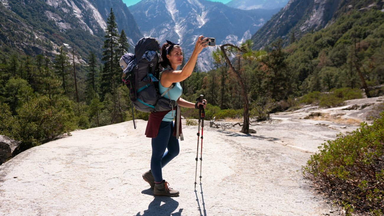 A hiker with a large backpack takes a photo with a smartphone against a stunning mountainous landscape and lush greenery.