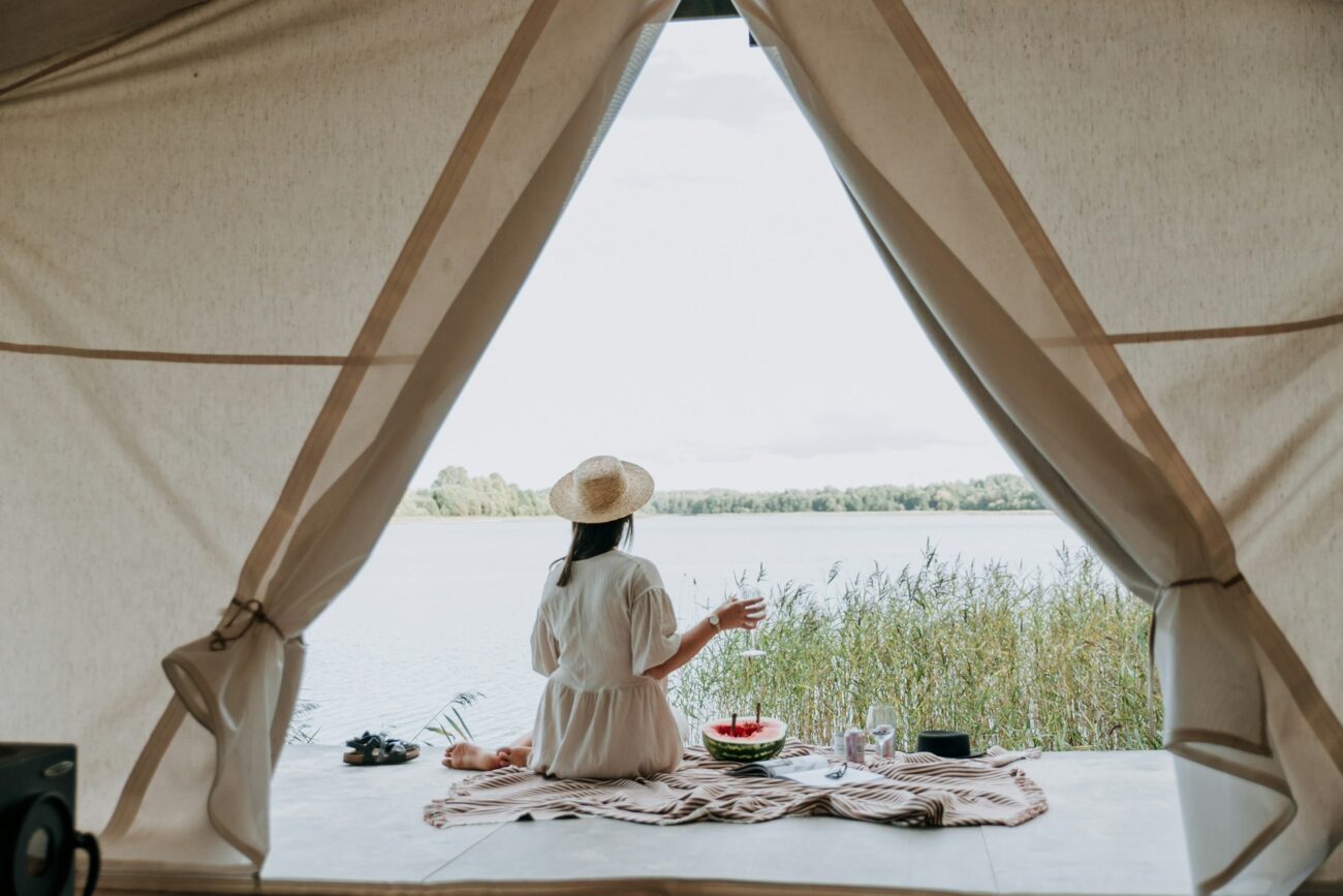 A woman in a white dress and straw hat sits by the water, enjoying refreshments and the serene view through a tent opening.