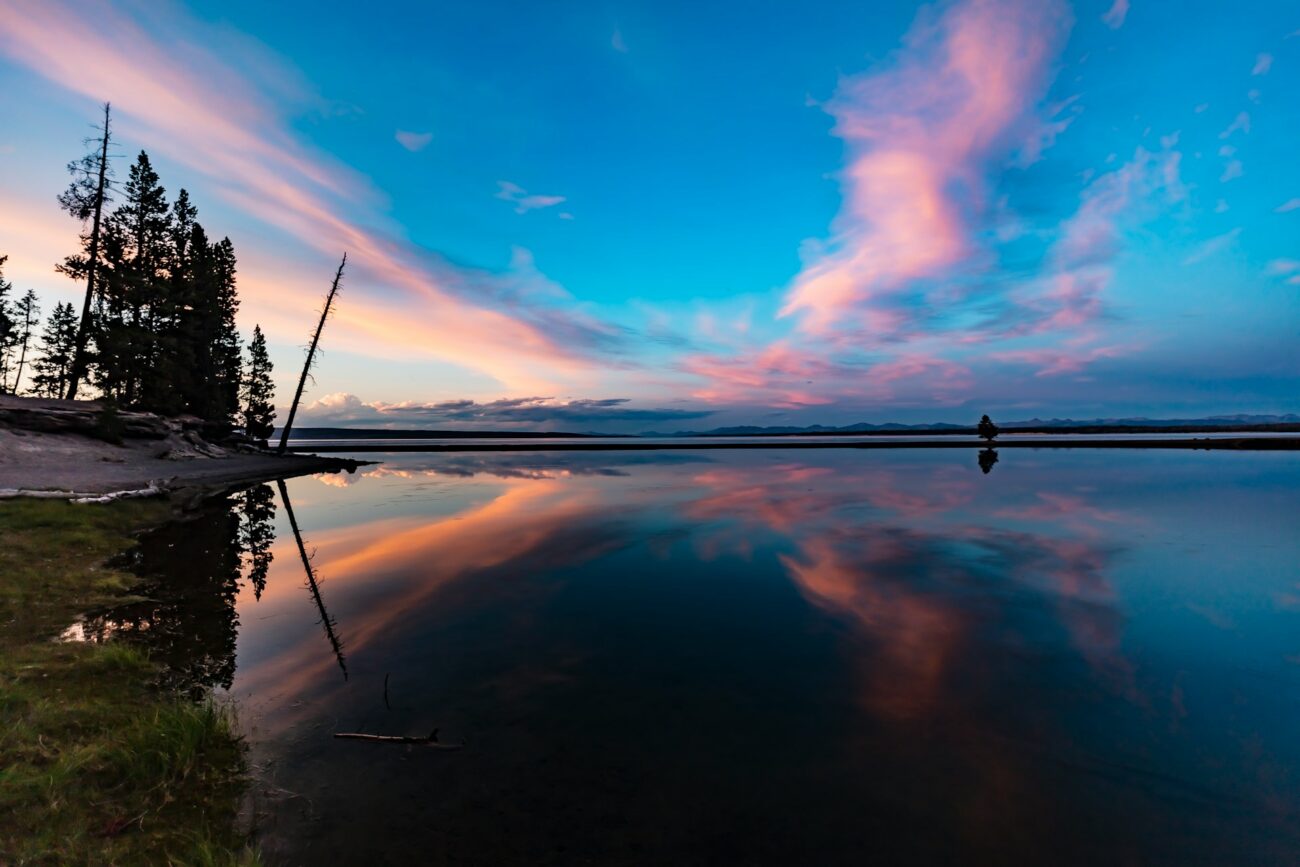 Tranquil sunset over a lake with vibrant pink and blue clouds reflecting on the water, surrounded by silhouetted trees.