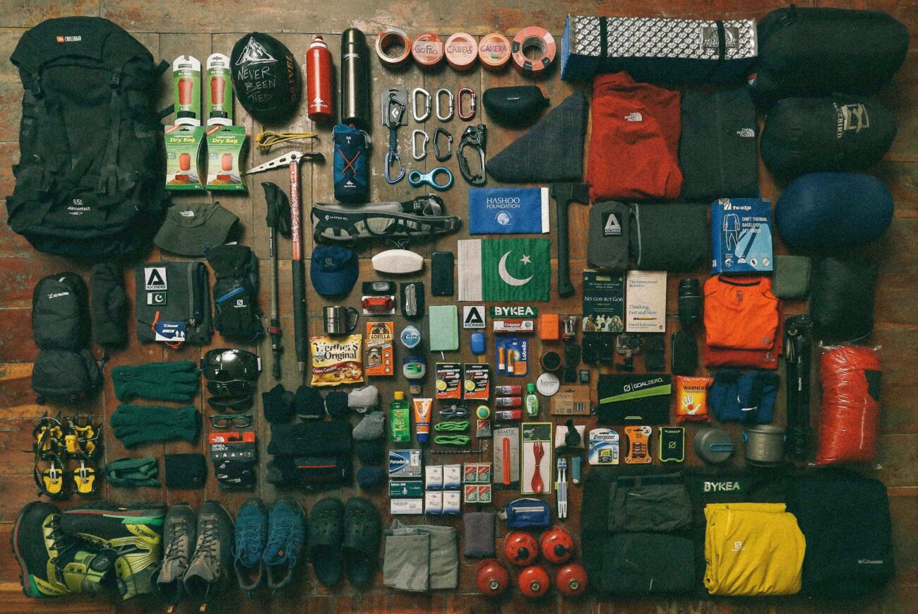 A flat lay of various outdoor gear including backpacks, clothing, snacks, and tools organized neatly on a wooden surface.