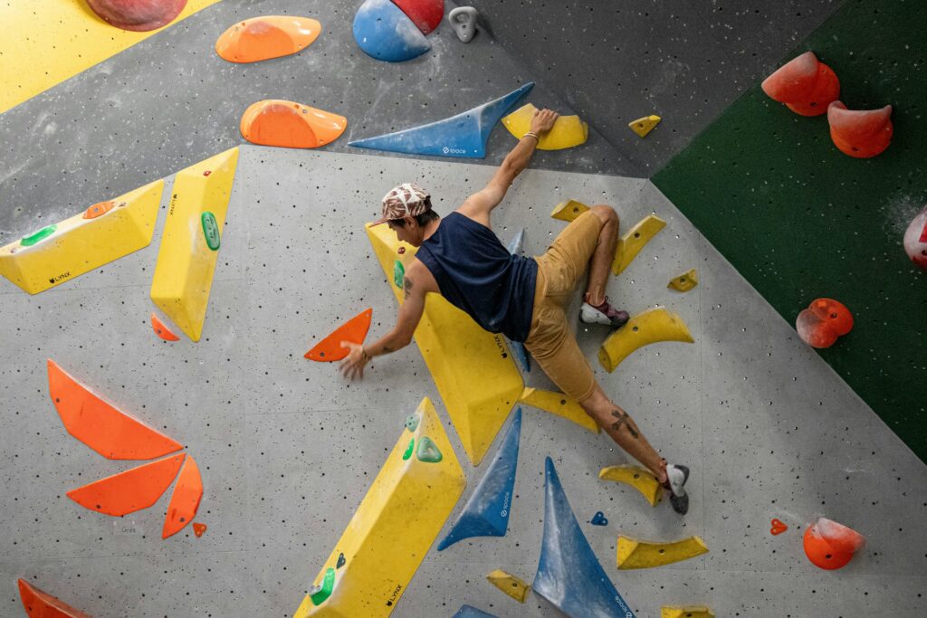 A climber maneuvers on a colorful indoor climbing wall, using vibrant holds in yellow, orange, and blue.