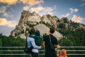 A family stands on a viewpoint overlooking a mountain landscape with lush greenery and a dramatic sky at sunset.