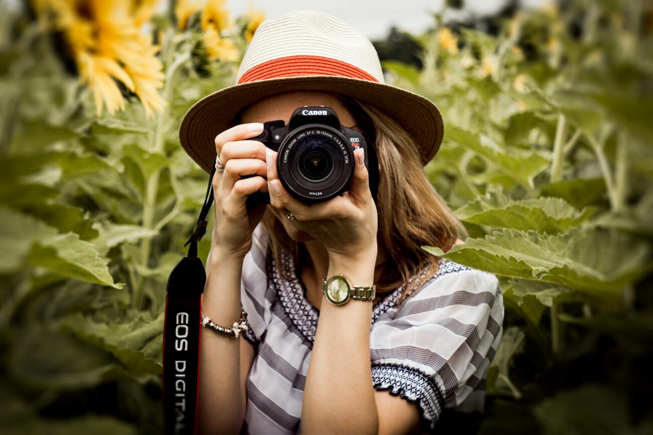 A woman in a hat holds a camera while standing in a vibrant field of sunflowers under a clear blue sky.