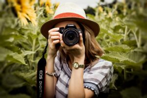 A woman in a hat holds a camera while standing in a vibrant field of sunflowers under a clear blue sky.