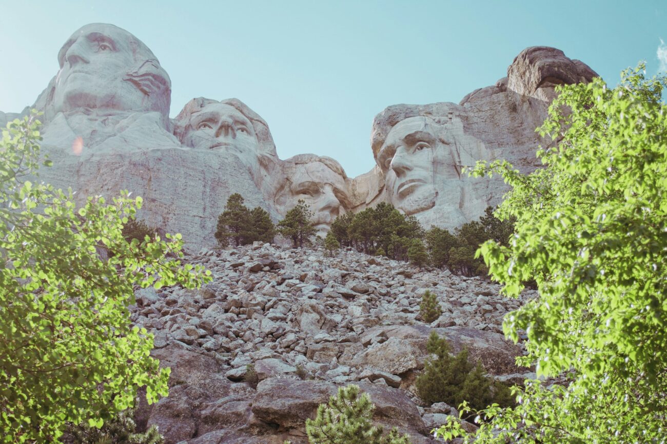 A rocky hillside dotted with lush green trees under a bright blue sky, framed by majestic mountain sculptures in the background.