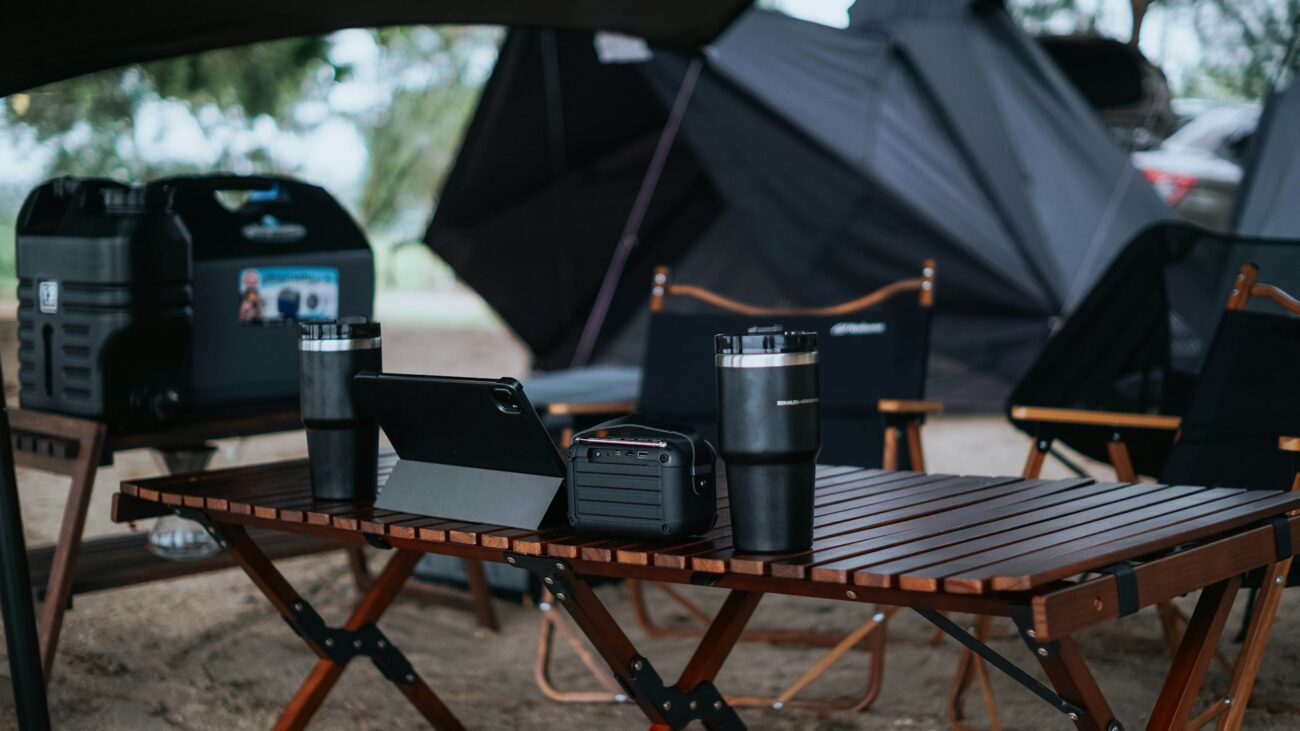 A cozy camping setup featuring a wooden table with two mugs, a tablet, and a portable battery, surrounded by dark tents.