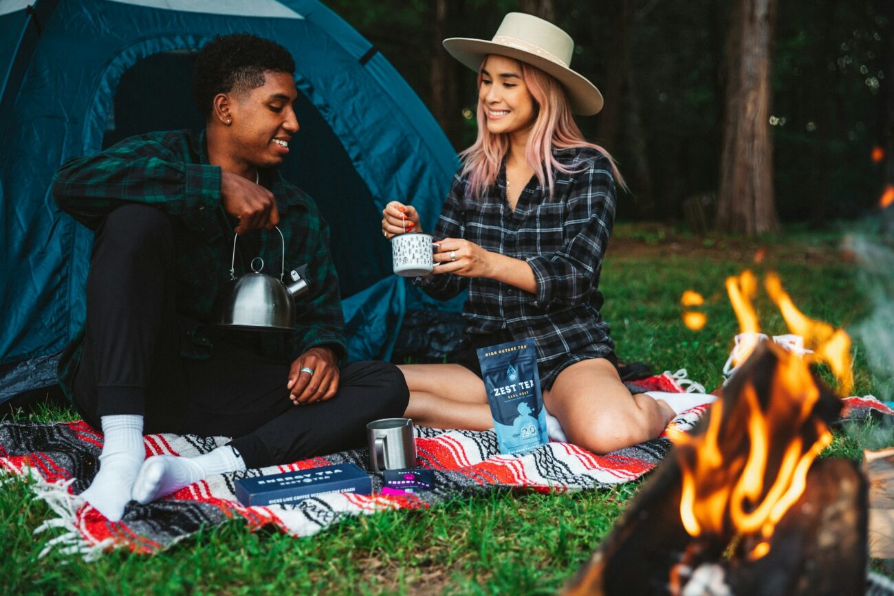 A couple enjoying tea by a campfire, seated on a blanket, with a tent in the background and a kettle in use.